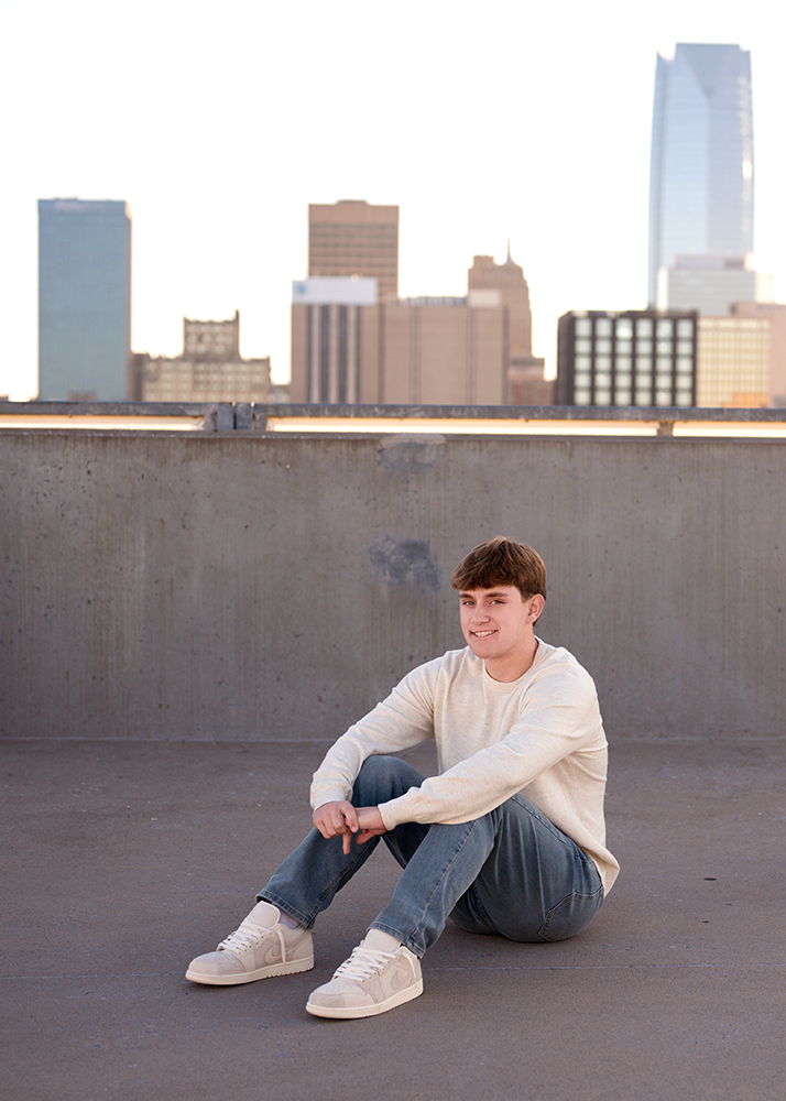 High school senior in a white sweater and jeans sits on a rooftop, smiling. The Oklahoma City skyline with skyscrapers towers in the background under a clear sky.