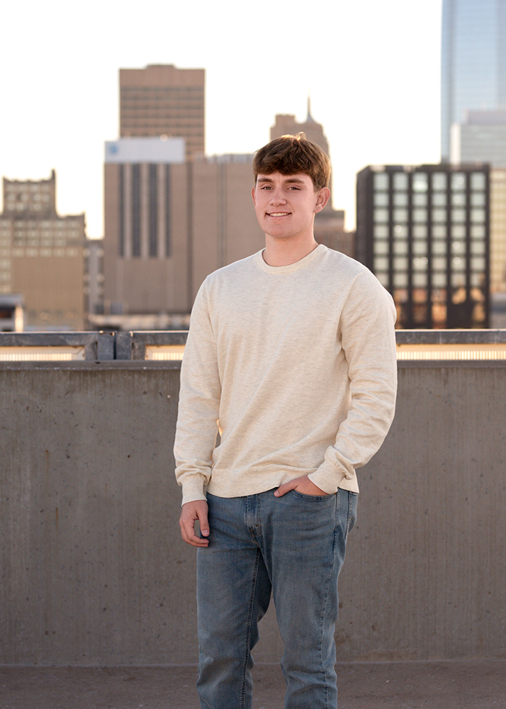 Senior guy in a white sweater and jeans sits on a rooftop, smiling. The Oklahoma City skyline with skyscrapers towers in the background under a clear sky.