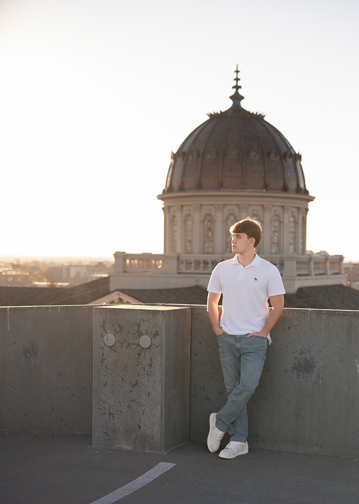 A high school senior in a white polo and jeans leans against a concrete wall on a rooftop at sunset. A domed building is in the background, creating a serene, reflective mood.