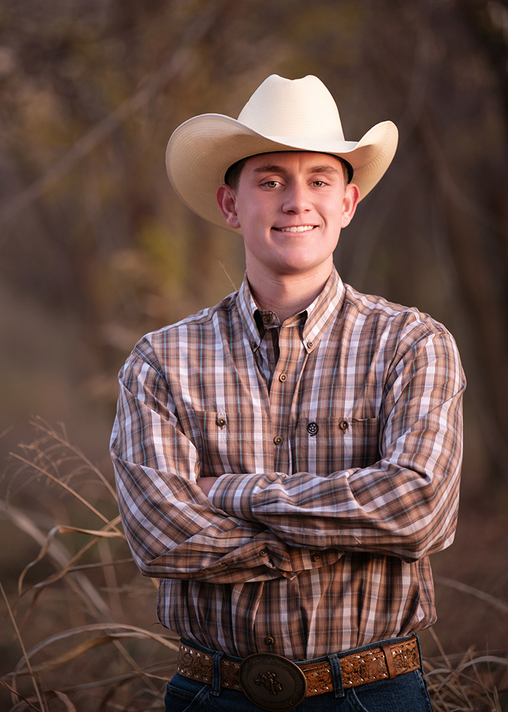 Young man in a plaid shirt and cowboy hat standing outdoors with arms crossed, smiling confidently. The background is soft-focused, suggesting a natural setting.