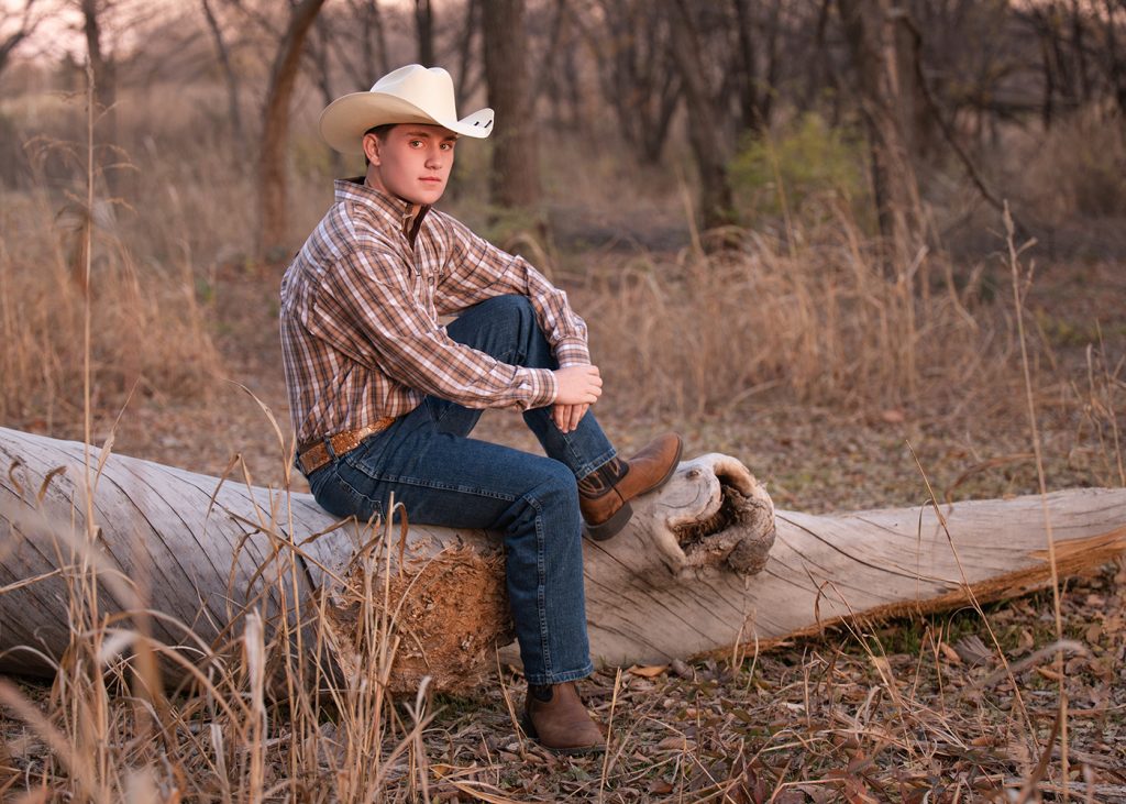 William wears a cowboy hat sits on a fallen log in a serene forest, wearing a plaid shirt, jeans, and boots. The setting sun casts a warm glow.