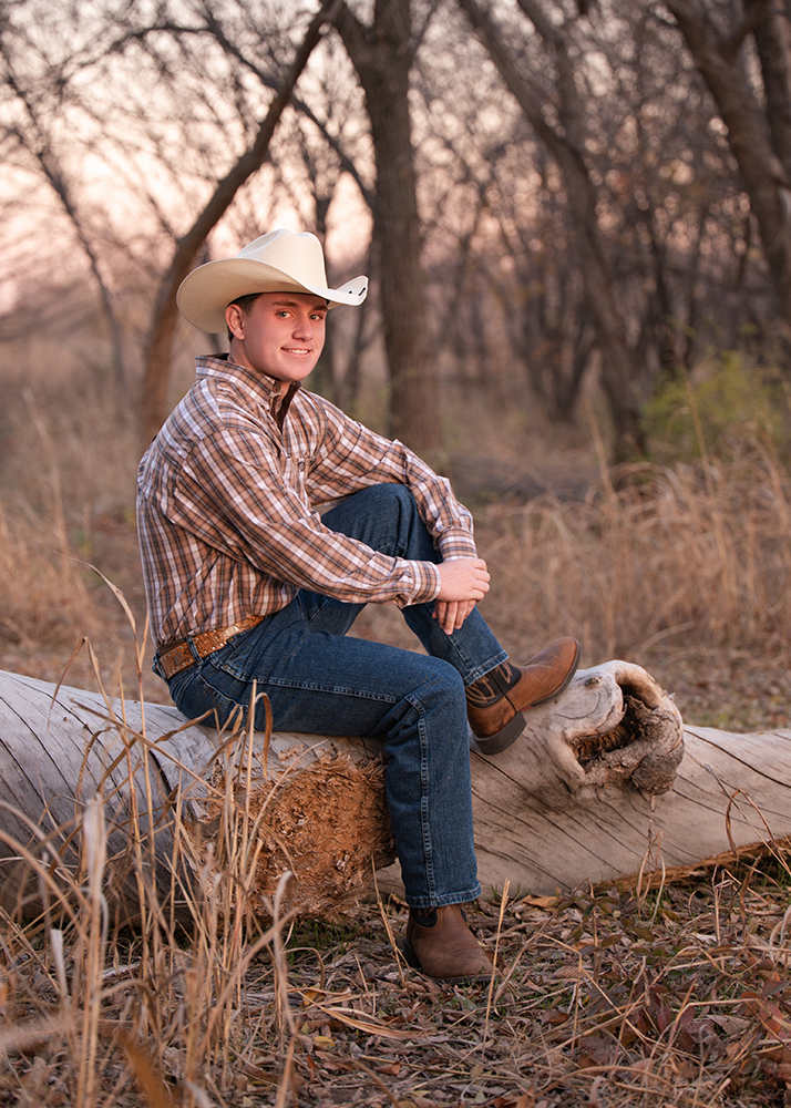 High school senior wears a cowboy hat sits on a fallen log in a serene forest, wearing a plaid shirt, jeans, and boots. The setting sun casts a warm glow.