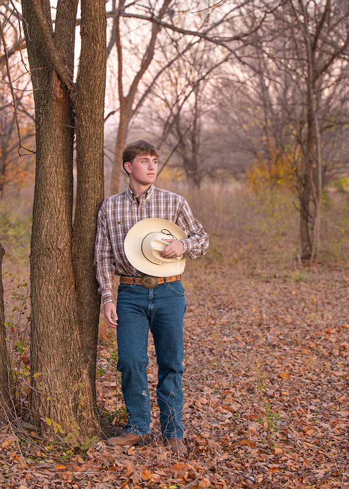 A young man in a plaid shirt and jeans leans against a tree, holding a cowboy hat. The autumn forest background creates a serene, contemplative mood.