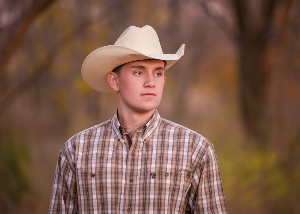 Senior photos of a young man in a cowboy hat and plaid shirt gazes thoughtfully into the distance. Soft, blurred autumnal background adds warmth and calmness to the scene.