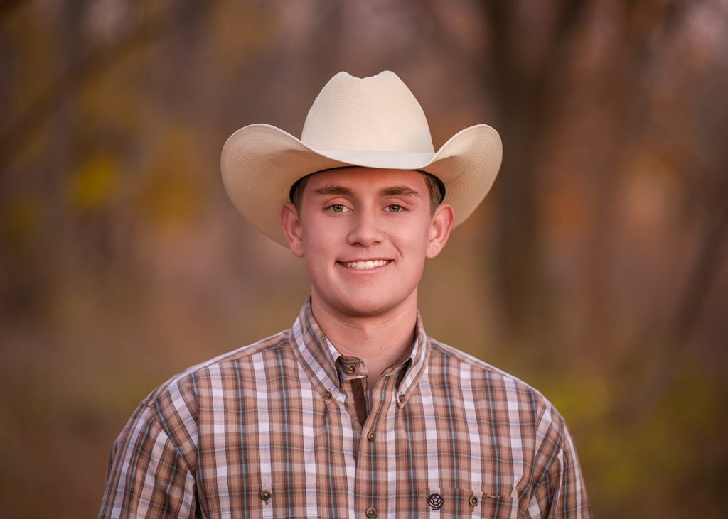 Photo session of a senior guy in a tan cowboy hat and plaid shirt stands outdoors, with a blurred autumnal background, conveying a warm, rustic feel.