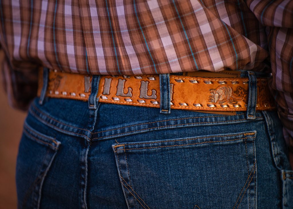 Close-up of a person wearing blue jeans and a brown plaid shirt. The tan leather belt features embossed designs and a name in bold letters.