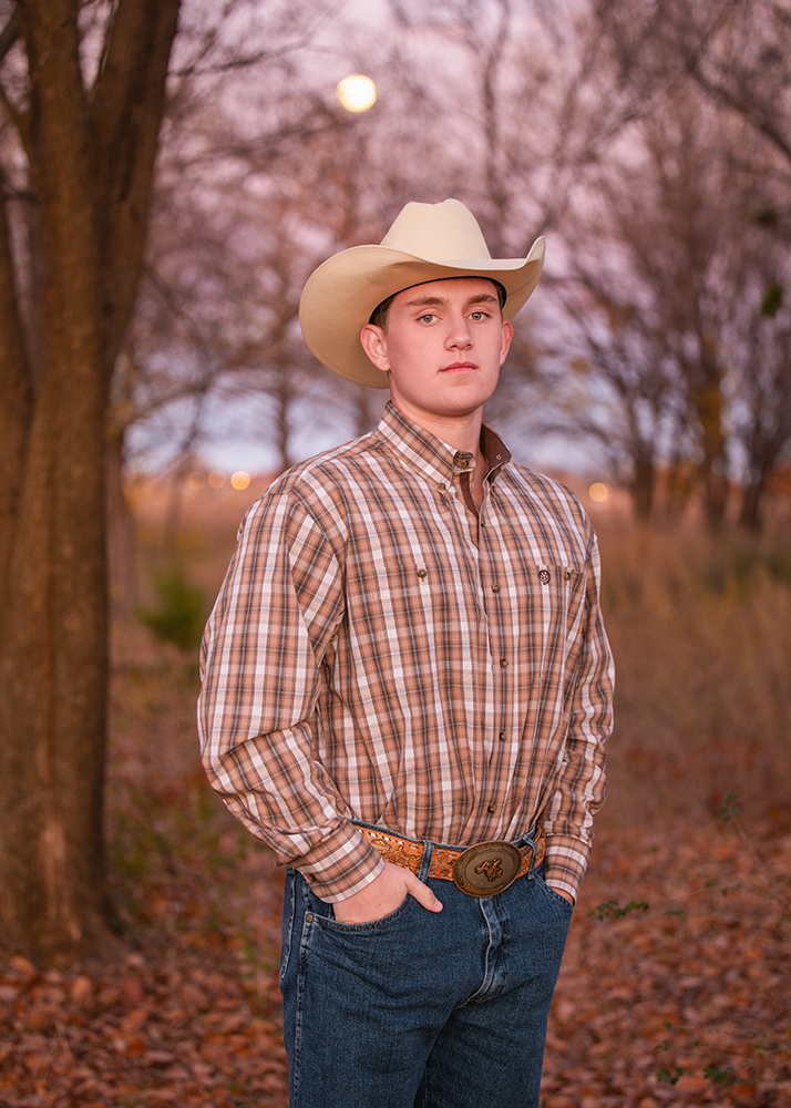High school senior takes senior photos in a cowboy hat and plaid shirt stands confidently in a serene autumn forest, with a soft pink and purple sunset sky as the backdrop.