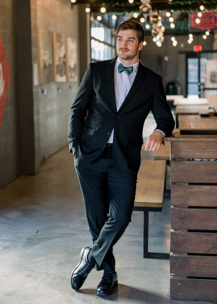 High school senior boy in black suit and bowtie posing indoors with modern industrial lighting