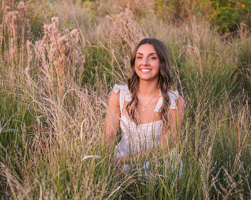 High school senior girl sitting in tall grass field during Oklahoma City senior portrait session with natural light