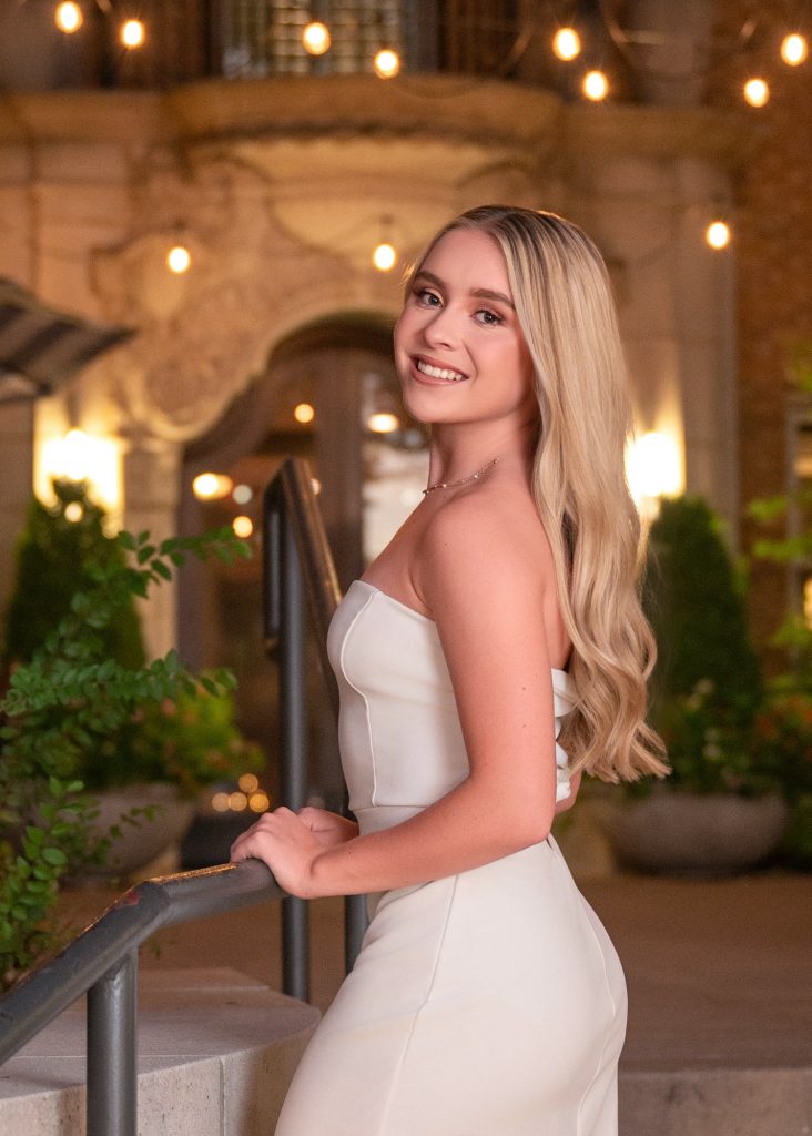 High school senior girl in elegant white dress posing at sunset with warm lights and greenery