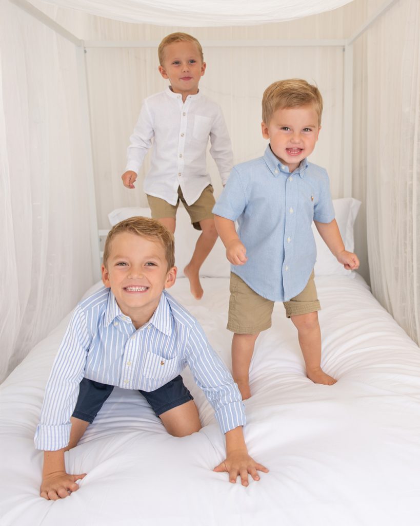 Three young brothers playing and laughing on a bed during a bright studio family photography session in Edmond, Oklahoma.